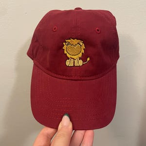May include: A burgundy baseball cap with a curved brim. The cap features an embroidered cartoon lion in yellow and tan on the front. The cap is made of a soft fabric and has a classic six-panel design.