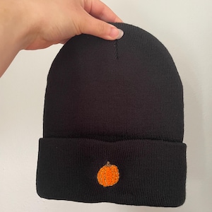 Puede incluir: Gorro de punto negro con borde doblado. Una calabaza naranja bordada está centrada en el borde doblado. El gorro está sujeto por una mano sobre un fondo blanco. Un accesorio con temática otoñal.