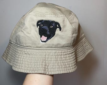 Puede incluir: Un sombrero de pescador beige con un parche bordado de una cara de perro negra con una lengua rosa. El sombrero tiene una copa redondeada y un ala inclinada hacia abajo con detalles de costuras visibles alrededor de la estructura.