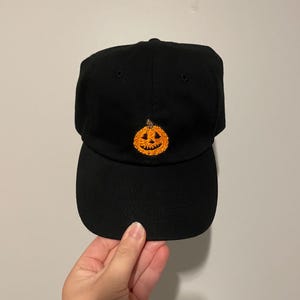 May include: Black baseball cap with an embroidered orange pumpkin design. The cap has a curved brim and a classic six-panel construction. A festive accessory for Halloween or autumn.