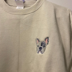 Puede incluir: Una sudadera beige con un bulldog francés bordado en la parte delantera.