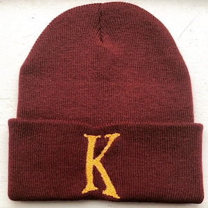 Op de afbeelding: Een bordeauxrode beanie met een gele geborduurde letter K aan de voorkant.