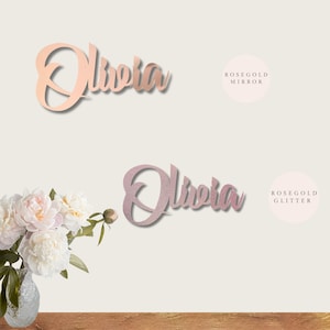 May include: Two versions of the name "Olivia" in rose gold. The top version is a rose gold mirror finish. The bottom version is a rose gold glitter finish.
