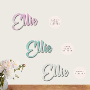 May include: Three versions of the name "Ellie" in cursive script, each with a different glitter finish: candy floss, sage green, and white.