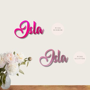 May include: Two versions of the name "Isla" in pink. The top version is a pink mirror finish. The bottom version is a pink glitter finish.