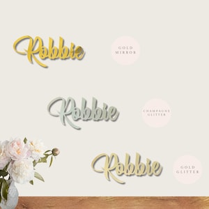 May include: Three versions of the name "Robbie" in different finishes: gold mirror, champagne glitter, and gold glitter. The name is written in a cursive script font.