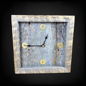May include: Square wooden wall clock with a rustic, weathered gray finish. The clock face is made of reclaimed wood, accented with brass bullet casings and copper rivets. Black clock hands indicate the time.
