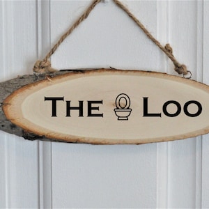 Può includere: Un cartello di legno con il testo "THE loo" e un'icona del water. Il cartello è di forma ovale e ha un aspetto rustico. È appeso a una corda.