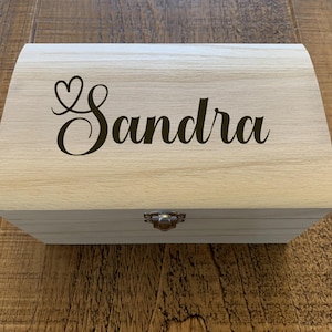 Puede incluir: Una caja de madera con tapa abatible y cierre de metal. La caja está decorada con el nombre "Sandra" y un diseño de corazón.
