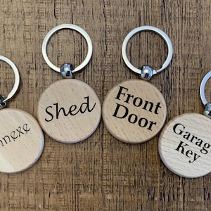 May include: Four wooden keychains with silver rings. Each keychain has a round wooden tag with black text: "Annexe", "Shed", "Front Door", and "Garage Key". The keychains are on a brown wooden surface.