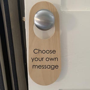 Op de afbeelding: Houten deurhanger met een zilveren knop en de tekst "Kies je eigen bericht".