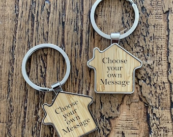 May include: Two house-shaped keychains with silver key rings. The keychains are made of light-colored wood and feature the text "Choose your own Message" in black. The keychains are displayed on a wooden surface.