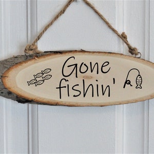 Może przedstawiać: Drewniany znak z napisem "Gone fishin'" i rysunkiem wędki i ryby. Nad tekstem pływa też kilka ryb w rzędzie.