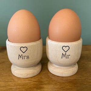 Peut inclure: Deux supports à œufs en bois avec un cœur et les mots "Mr" et "Mrs" dessus. Les supports à œufs contiennent des œufs bruns.