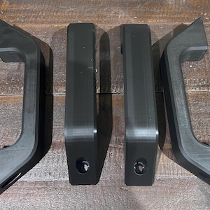 May include: Four black, angular handles are arranged on a wooden surface. Two handles have a curved design, while the other two are rectangular. Each handle has a hole. The handles appear to be made of a matte material.