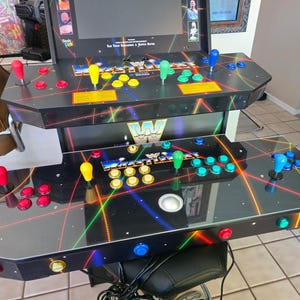 May include: A black tabletop arcade game console featuring WWE WrestleFest branding, a screen, and two control panels. Each panel includes a joystick and multiple colored buttons in red, yellow, green, and blue, set against a neon laser graphic.