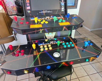May include: A black tabletop arcade game console featuring WWE WrestleFest branding, a screen, and two control panels. Each panel includes a joystick and multiple colored buttons in red, yellow, green, and blue, set against a neon laser graphic.