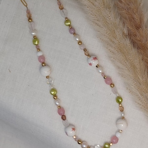 Puede incluir: Un delicado collar de cadena dorada con cuentas rosas, verdes, blancas y transparentes. Las cuentas tienen un patrón floral y están intercaladas con detalles dorados.