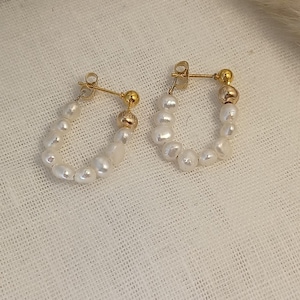 Puede incluir: Un par de pendientes de aro dorados con cierre de poste. Cada pendiente está adornado con una serie de pequeñas perlas blancas irregulares. Una pequeña cuenta dorada está colocada en la parte superior de cada pendiente.