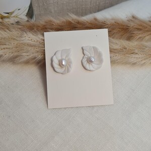 Puede incluir: Un par de pendientes blancos con forma de concha, cada uno con una pequeña perla redonda en el centro. Se exhiben sobre una tarjeta color crema, sobre un fondo neutro con hierbas secas.