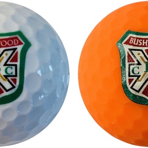 May include: Two golf balls, one white and one orange, each with the "Bushwood CBC" logo. The logo is a shield design with the text "Bushwood" at the top, and "CBC" at the bottom, with crossed golf clubs.