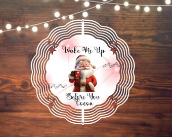 May include: A circular Christmas-themed design featuring Santa Claus holding a mug of cocoa. The text "Wake Me Up Before You Cocoa" is displayed. The design has a white scalloped edge with red stars and musical notes.