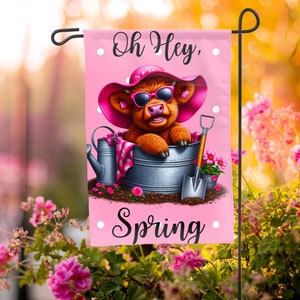 May include: A pink garden flag with a cartoon Highland cow wearing sunglasses and a pink hat. The cow is sitting in a metal bucket with a watering can and a shovel. The text on the flag reads "Oh Hey, Spring".