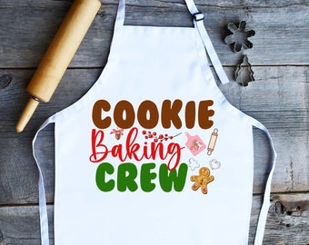 Könnte beinhalten: Weiße Schürze mit dem Text "COOKIE Baking CREW" in braunen, roten und grünen Buchstaben. Die Schürze zeigt einen Lebkuchenmann, einen Nudelholz, einen Ausstecher und einen Zweig Stechpalme.
