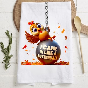 May include: A white kitchen towel with a cartoon chicken holding onto a weighted ball that says "I came in like a butterball". The chicken is brown and yellow with a red comb and wattle. The ball is gray and has a chain attached to it. There are orange, yellow, and brown leaves scattered around the chicken and ball.