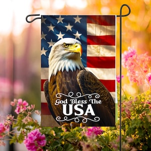 May include: A garden flag featuring an American flag design with an eagle and the text "God Bless The USA." The flag is displayed on a black metal stand, surrounded by pink flowers and greenery.