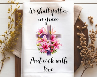 May include: White kitchen towel with a purple cross and pink lilies. The text "We shall gather in grace and cook with love" is printed on the towel.