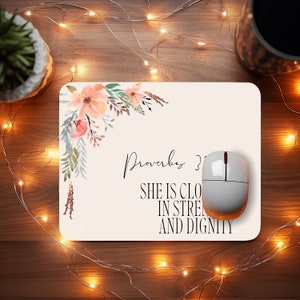 Op de afbeelding: Een wit muismatje met een bloemmotief in de linkerbovenhoek. De tekst "Spreuken 31" en "Zij is bekleed met kracht en waardigheid" staat in het zwart op het muismatje.