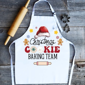 May include: White Christmas cookie baking apron featuring a red Santa hat, gingerbread men, and a rolling pin graphic. The text reads "Christmas Cookie Baking Team".