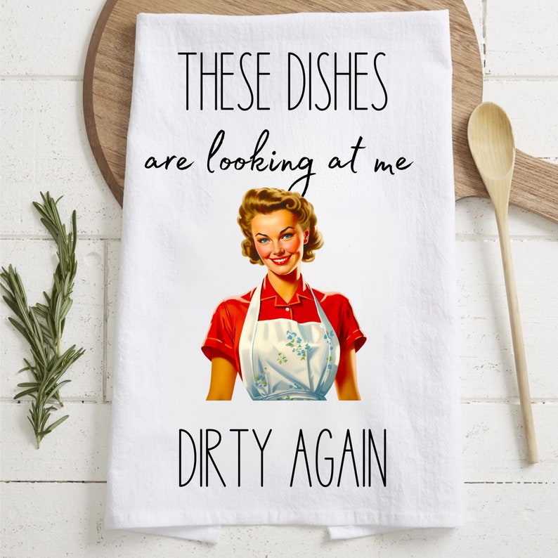 May include: White kitchen towel with a retro illustration of a woman in a red and white apron. The text reads "These dishes are looking at me dirty again."