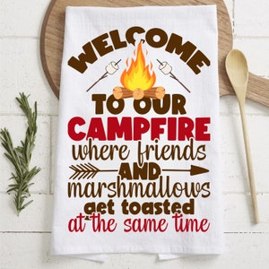 May include: White kitchen towel with brown text that reads "Welcome to our campfire where friends and marshmallows get toasted at the same time". The text is surrounded by a campfire scene with orange flames, brown logs, and white marshmallows on sticks.