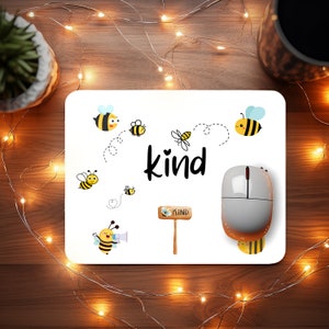 May include: White mouse pad with a bee theme. The mouse pad has a black and white bee illustration with the word "kind" in black text. There are several other bee illustrations on the mouse pad, including a bee holding a sign that says "kind".