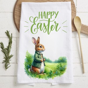 Könnte beinhalten: Ein weißes Küchentuch mit einer grünen Aufschrift "Frohe Ostern" und einer Cartoon-Illustration eines braunen Kaninchens, das eine grüne Jacke in einem Feld mit grünem Gras trägt.