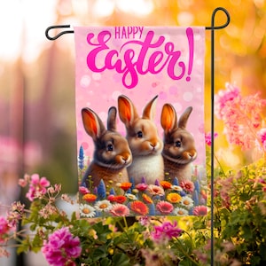 May include: A pink garden flag with the text "Happy Easter!" in pink script. Three brown and white bunnies are sitting in a field of colourful flowers.