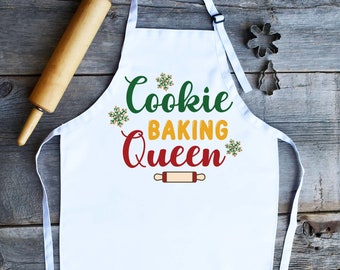 Könnte beinhalten: Weiße Schürze mit dem Text "Cookie Baking Queen" in grüner, gelber und roter Schrift. Unter dem Text befindet sich eine kleine Grafik eines Nudelholzes. Das Design ist mit Schneeflocken verziert.