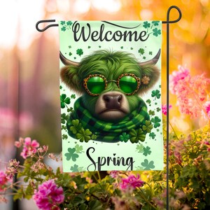 May include: A garden flag featuring a green Highland cow wearing sunglasses and a scarf, with the words "Welcome Spring." The flag is decorated with shamrocks and is displayed on a black metal stand. The background is blurred with pink flowers.