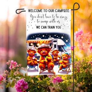 May include: A white garden flag with a cartoon illustration of four Highland cows sitting around a camper van. The text on the flag reads "WELCOME TO OUR CAMPSITE You don't have to be crazy to camp with us WE CAN TRAIN YOU".