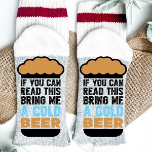 May include: A pair of white socks with red stripes at the top. The socks have a gray and white design on the bottom with a brown beer mug graphic and the text "IF YOU CAN READ THIS BRING ME A COLD BEER".