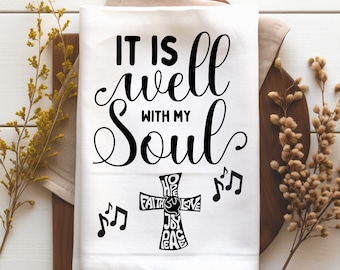 May include: White kitchen towel with black text that reads "It is well with my Soul". A black cross with the words "Hope, Faith, Joy, Love, Peace" is below the text. Musical notes are on either side of the cross.