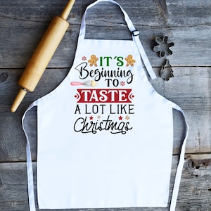 May include: White kitchen apron with a red and black graphic that says "It's Beginning to Taste a Lot Like Christmas". The graphic includes gingerbread men, snowflakes, and a whisk.