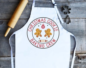 Könnte beinhalten: Weiße Schürze mit einem schwarzen Kreisdesign, auf dem in roter Schrift "Christmas Cookie Baking Crew" steht. Zwei Lebkuchenmänner befinden sich im Kreis, zusammen mit einem Nudelholz und einem roten Ornament.