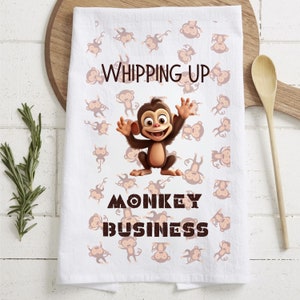 Puede incluir: Toalla de cocina blanca con un mono de dibujos animados marrón y el texto "Whipping Up Monkey Business" impreso en ella. La toalla está cubierta con un patrón de monos de dibujos animados marrones más pequeños.