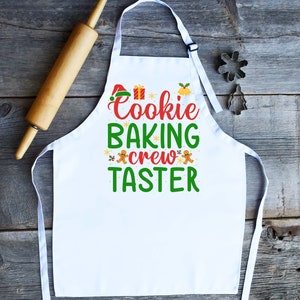 May include: White Christmas-themed apron with the text "Cookie Baking Crew Taster" in green and red lettering. The apron features gingerbread men, snowflakes, and a Santa hat.