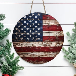Op de afbeelding: Een rond houten bord met een verweerd Amerikaans vlagontwerp. Het bord hangt aan een witte muur met twee groene dennentakken aan weerszijden.