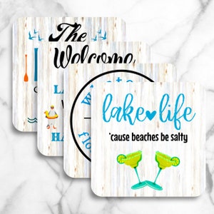 May include: Four wooden coasters with a whitewashed wood background and blue and black text. The coasters feature lake life themed sayings, including "The Welcome", "Lake Life 'cause beaches be salty", and "Welcome to the Lake".