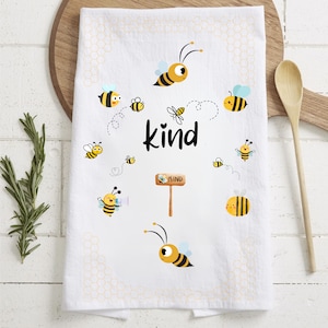 May include: White kitchen towel with a honeycomb pattern and multiple cartoon bees. The text "kind" is written in black letters. A small sign with the word "kind" is also on the towel.